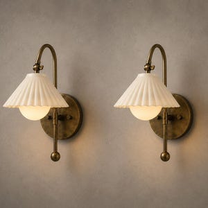 May include: A pair of wall sconces featuring a circular antique brass backplate, a curved arm, and a pleated off-white ceramic shade. Each fixture includes a central frosted glass globe bulb that emits a soft, warm light from beneath the shade.