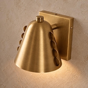 Pode incluir: Um candeeiro de parede com acabamento em latão escovado. A lâmpada apresenta um abajur cónico adornado com uma fila vertical de discos metálicos circulares na lateral. Uma placa quadrada liga o candeeiro à parede e um pequeno botão cilíndrico situa-se no topo do abajur.