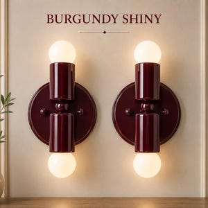 Könnte beinhalten: Zwei Wandleuchten mit glänzend burgunderroter Oberfläche, jeweils mit einer runden Rückplatte und zwei zylindrischen Fassungen, die kugelförmige weiße Glühbirnen halten. Der Text BURGUNDY SHINY ist mittig über den symmetrisch montierten Leuchten.