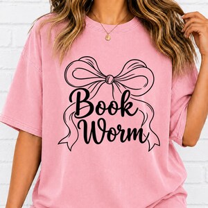 May include: A pink short-sleeved t-shirt featuring a black graphic of a large decorative bow centered on the chest. Below the bow, the text "Book Worm" is printed in a bold, black cursive font. The shirt has a crew neckline and a relaxed fit.