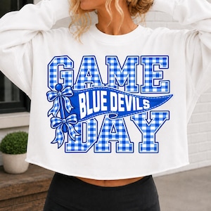 May include: A white long-sleeve cropped sweatshirt featuring a blue and white gingham pattern graphic. The text reads GAME DAY with a central blue pennant banner displaying the words BLUE DEVILS, accented by two gingham bows on the left side.