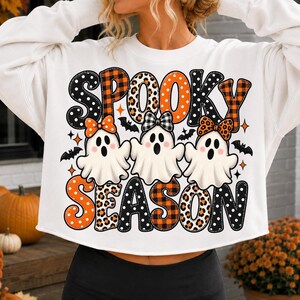 May include: A white long-sleeve sweatshirt featuring the text "SPOOKY SEASON" in large, patterned letters. Three white ghosts with surprised expressions and colorful bows are positioned between the words, surrounded by small black bats and stars.