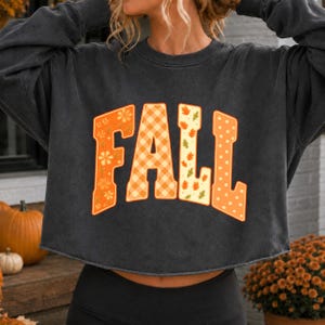 Könnte beinhalten: Ein anthrazitfarbenes Langarm-Sweatshirt mit Rundhalsausschnitt und offenem Saum. Auf der Vorderseite steht das Wort FALL in großen Blockbuchstaben, die jeweils ein einzigartiges orange-cremefarbenes Muster mit Blumen, Karos, Blättern und Punkten aufweisen.
