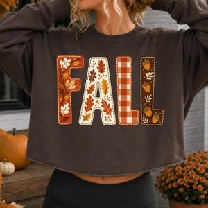 May include: A dark brown long-sleeve cropped sweatshirt featuring the word FALL printed in large block letters. Each letter has a unique pattern: the F has orange leaves, the A has autumn foliage, the L has a red plaid, and the L has acorns.