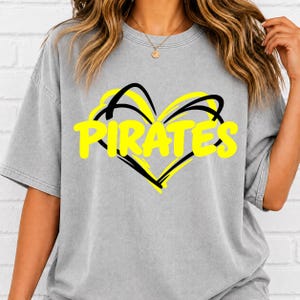 May include: A grey short-sleeve t-shirt featuring a graphic design of a black outlined heart shape with the word PIRATES printed in bold yellow capital letters across the centre of the heart. The shirt has a crew neckline and a relaxed fit.