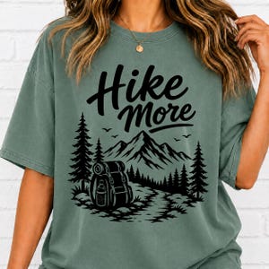 May include: A sage green short-sleeve t-shirt featuring a black graphic print of a backpack, pine trees, and mountains with the text "Hike More" in a bold script font. The shirt has a crew neckline and a relaxed, loose-fitting silhouette.