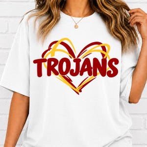 May include: A white short-sleeved t-shirt featuring a graphic design of a heart outlined in yellow and red brushstrokes. The word TROJANS is printed in bold, dark red, rounded capital letters across the center of the heart graphic.