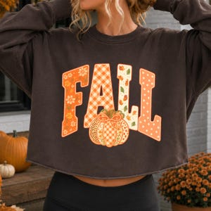 May include: A dark brown long-sleeve cropped sweatshirt featuring the word FALL in large block letters. Each letter has a unique pattern including orange flowers, white and orange gingham, autumn leaves, and white polka dots on an orange background.