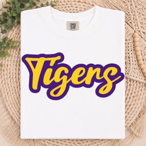May include: A white short-sleeved t-shirt featuring the word Tigers printed in a bold yellow script font with a thick purple outline. The shirt has a Comfort Colors brand label at the inner neckline and a classic crew neck design.