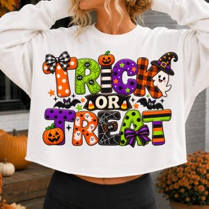 May include: A white long-sleeve sweatshirt featuring a colorful graphic with the text Trick or Treat. The design includes pumpkins, ghosts, bats, spiders, spiderwebs, bows, and a witch hat in orange, green, purple, and black patterns.