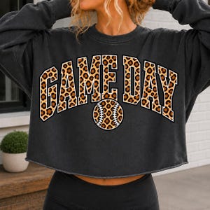 Puede incluir: Una sudadera corta de manga larga en color gris carbón con el texto GAME DAY en una fuente universitaria arqueada y llamativa. Las letras y un gráfico central de béisbol están rellenos con un estampado de leopardo marrón y negro.