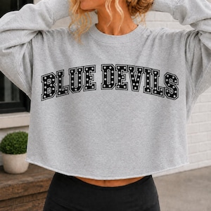 May include: A light grey cropped sweatshirt featuring long sleeves and a crew neckline. The front displays the text "BLUE DEVILS" in a bold, black collegiate-style font with a white polka dot pattern inside each letter and a black outline.