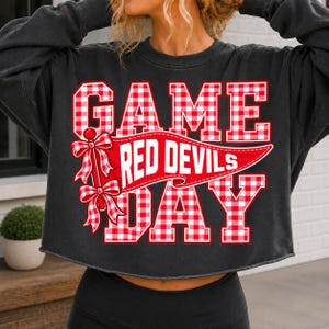 May include: A charcoal gray long-sleeve cropped sweatshirt featuring a graphic print with the text GAME DAY in a red and white gingham pattern. A red and white gingham pennant banner with the text RED DEVILS and two bows is centered.