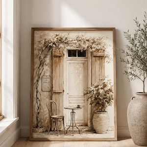 May include: A framed wall art print depicting a beige and brown scene of a wooden door with shutters, a small bistro chair, a round table with a cup, and a potted plant. A climbing vine hangs above the door, and a sign reads Maison de Famille.