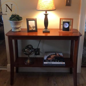 May include: A wooden console table with a top surface holding a lamp with a textured shade, a framed photograph, and a square clock. The lower shelf features a metal armillary sphere and a stack of three books titled Photo Ark and Hopper.