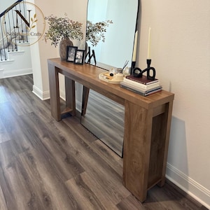 May include: A rectangular wooden console table with a natural brown finish and thick legs. The surface holds a round tray with small items, a stack of books, two black candle holders with white taper candles, two framed photos, and a vase with greenery.