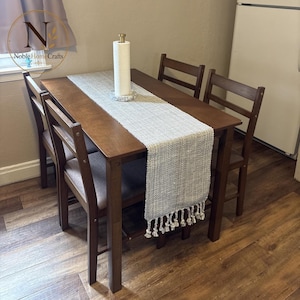 May include: A rectangular wooden dining table with four matching chairs, featuring a light gray woven table runner with fringe ends. A white paper towel roll sits on a circular marble base in the center of the table, which is set on a wood floor.