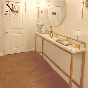 May include: A tall, narrow console table with a white top and a gold metal frame. The table holds a white vase with flowers, a white jar, a gold abstract sculpture, a gold tray with a white candle, and a small gold lamp with a pleated shade.