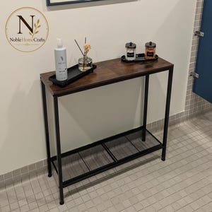 May include: A rectangular console table with a dark brown wood-grain top and a black metal frame. The table features a lower shelf made of black wire mesh. On the top surface, there is a white lotion bottle, a reed diffuser, and two glass jars.