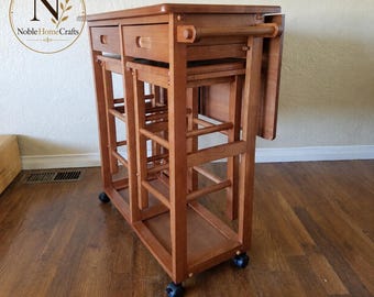 May include: A brown wooden kitchen island cart featuring a rectangular top, a drawer, and a side handle. The cart includes a matching stool tucked underneath and is supported by four black caster wheels for mobility on the floor.