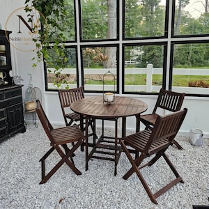 May include: A dark brown wooden patio set consisting of a round slatted table and four matching folding chairs. The furniture is arranged on a white gravel surface. A small metal lantern sits in the center of the table, and a green plant hangs.