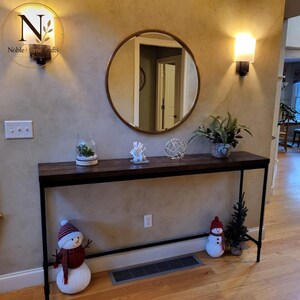 May include: A long, narrow console table with a dark wood top and a black metal frame. The table is decorated with a glass jar, a white coral-like sculpture, a wire sphere, a potted plant, and two plush snowman figures on the floor.