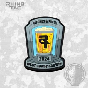 May include: A grey, shield-shaped morale patch featuring a central graphic of a beer glass filled with yellow liquid. The text reads PATCHES & PINTS, 2024, and WEST COAST EDITION. A black logo is centred on the glass against a light blue background.