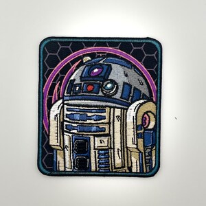 May include: A rectangular embroidered patch featuring a stylized blue, white, and silver robot character. The design includes a dark background with a subtle hexagonal pattern and a vibrant purple circular arc framing the top of the robot figure.
