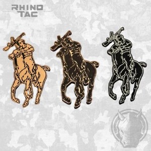 Può includere: Tre spille raffiguranti un cavaliere a cavallo che impugna un fucile, presentate in marrone chiaro, marrone scuro e nero. Il testo RHINO TAC appare in alto a sinistra e un logo con testa di rinoceronte è visibile nell'angolo in basso a destra.