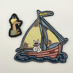 Peut inclure: Deux écussons brodés représentant des personnages de Max et les Maximonstres. L'un montre un monstre beige à cornes, et le plus grand représente un voilier sur des vagues bleues avec un personnage en costume de loup blanc et le nom MAX.
