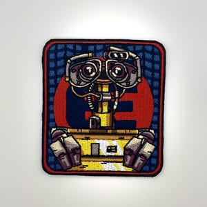 May include: A rectangular embroidered patch featuring a robot with large circular eyes, a yellow torso, and mechanical arms. The background consists of a red circle set against a blue grid pattern, all enclosed within a dark blue border.