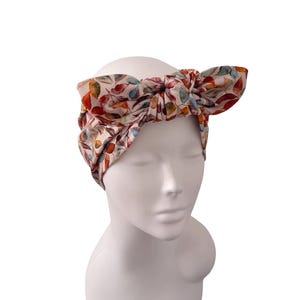 May include: A fabric headband featuring a knotted bow detail at the top. The headband is decorated with a repeating pattern of watercolour-style leaves in shades of orange, red, blue, and green against a light, off-white background colour.