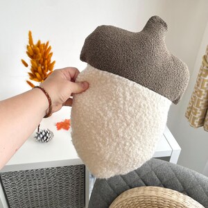 May include: An acorn-shaped decorative pillow featuring a textured brown bouclé fabric on the top cap section and a soft, cream-colored sherpa-style fleece material on the bottom nut portion, creating a distinct two-tone appearance.