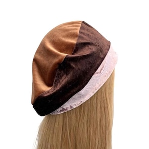 Op de afbeelding: Een meerkleurige muts met een glad beige bovendeel, een donkerbruine fluwelen middenband en een lichtroze textuurrand aan de onderkant. De muts heeft een afgerond, losvallend silhouet en is opgebouwd uit contrasterende stoffen panelen.