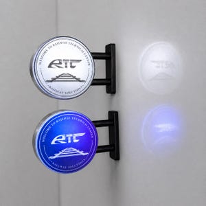 Könnte beinhalten: Zwei kreisförmige Wandleuchten mit schwarzen Metallhalterungen. Das obere Schild ist silberfarben mit schwarzer Aufschrift Welcome to Railway Technical Center und Railway Solutions, einem RTC-Logo und Gleisen. Das untere Schild leuchtet blau.