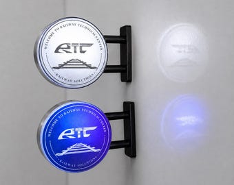 Könnte beinhalten: Zwei kreisförmige Wandleuchten mit schwarzen Metallhalterungen. Das obere Schild ist silberfarben mit schwarzer Aufschrift Welcome to Railway Technical Center und Railway Solutions, einem RTC-Logo und Gleisen. Das untere Schild leuchtet blau.