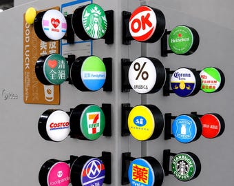 Könnte beinhalten: Eine Sammlung kleiner, runder, beleuchteter Schilder mit verschiedenen Markenlogos wie Starbucks, Heineken, Costco, 7-Eleven, FamilyMart und anderen. Jedes Schild ist an einer schwarzen Halterung auf einer hellgrauen Oberfläche befestigt.