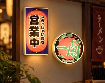 May include: A rectangular wall-mounted light box featuring a red lantern graphic with Japanese text and a circular light box displaying the Ichiran logo in green and red. Both signs are illuminated and mounted on a light-colored wall surface.