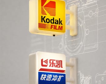 Könnte beinhalten: Zwei quadratische, beleuchtete Wandleuchtschilder. Das obere Schild zeigt das gelb-rote Kodak Film Logo. Das untere Schild zeigt rote und blaue chinesische Schriftzeichen mit einem grafischen Symbol. Beide haben weiße Rahmen und Halterungen.