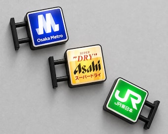 Könnte beinhalten: Drei quadratische, wandmontierte Schilder mit schwarzen Rahmen. Das obere Schild ist blau mit dem weißen Osaka Metro Logo und Schriftzug. Das mittlere Schild ist gelb-weiß mit dem Asahi Super Dry Logo und Schriftzug. Das untere Schild ist grün mit dem JR Logo.
