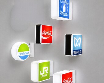 Könnte beinhalten: Eine Sammlung von sechs wandmontierten, beleuchteten quadratischen und runden Schildern mit verschiedenen Markenlogos wie Lawson Station, Coca-Cola, FamilyMart, Tokyo Metro und JR East, jeweils mit weißem Rahmen und Innenbeleuchtung.