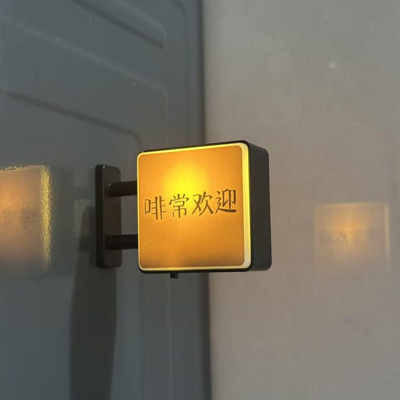 May include: A square, wall-mounted LED sign with a black frame and a glowing, warm yellow face. The sign features the Chinese characters 啡常欢迎 in black, centered on the illuminated surface, and is attached to the wall by a black bracket.