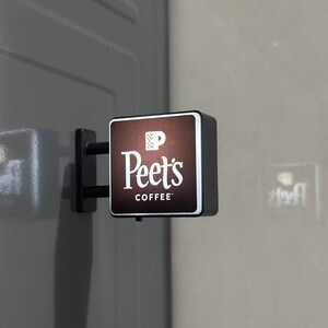 May include: A square, wall-mounted Peet's Coffee sign with a dark brown face, white lettering, and a white logo icon. The sign features a black frame and a black mounting bracket attached to a gray surface, with a small power switch on the bottom.