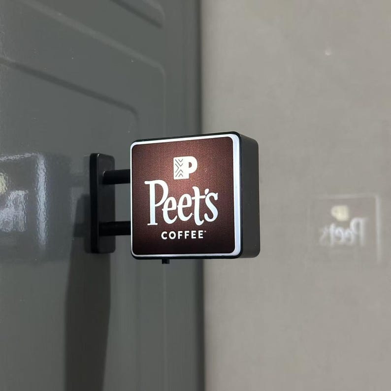 May include: A square, wall-mounted Peet's Coffee sign with a dark brown face, white lettering, and a white logo icon. The sign features a black frame and a black mounting bracket attached to a gray surface, with a small power switch on the bottom.