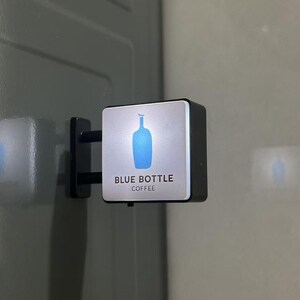 May include: A square, wall-mounted sign featuring a silver face with a blue bottle graphic and the black text "BLUE BOTTLE COFFEE" printed below. The sign has a black frame and is attached to a vertical surface by two black horizontal metal arms.