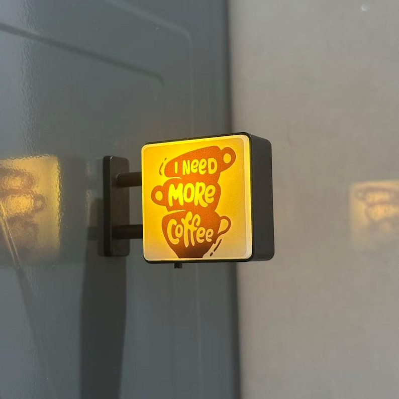 May include: A square, wall-mounted LED light box with a black frame and a warm yellow glow. The front panel features a graphic of three stacked coffee cups with the text "I need MORE Coffee" printed in a dark brown, stylized script font.