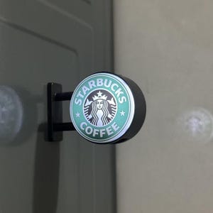 May include: A circular Starbucks Coffee sign featuring the iconic green and white siren logo. The sign is mounted on a black bracket with two horizontal bars, extending from a flat surface. The design includes the text Starbucks Coffee.