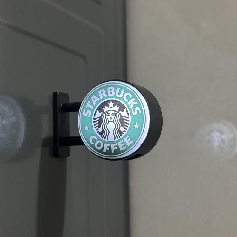 May include: A circular Starbucks Coffee sign featuring the iconic green and white siren logo. The sign is mounted on a black bracket with two horizontal bars, extending from a flat surface. The design includes the text Starbucks Coffee.
