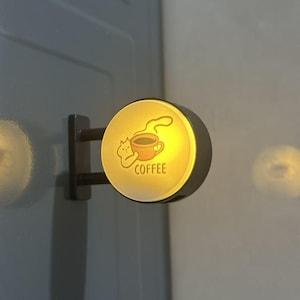 May include: A circular, wall-mounted LED sign with a warm yellow glow. The face features a graphic of a small cat holding a steaming mug of coffee, with the word COFFEE printed below in a simple, dark sans-serif font. The sign has a black frame.