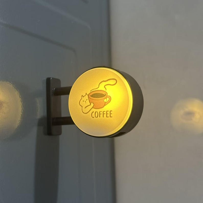 May include: A circular, wall-mounted LED sign with a warm yellow glow. The face features a graphic of a small cat holding a steaming mug of coffee, with the word COFFEE printed below in a simple, dark sans-serif font. The sign has a black frame.